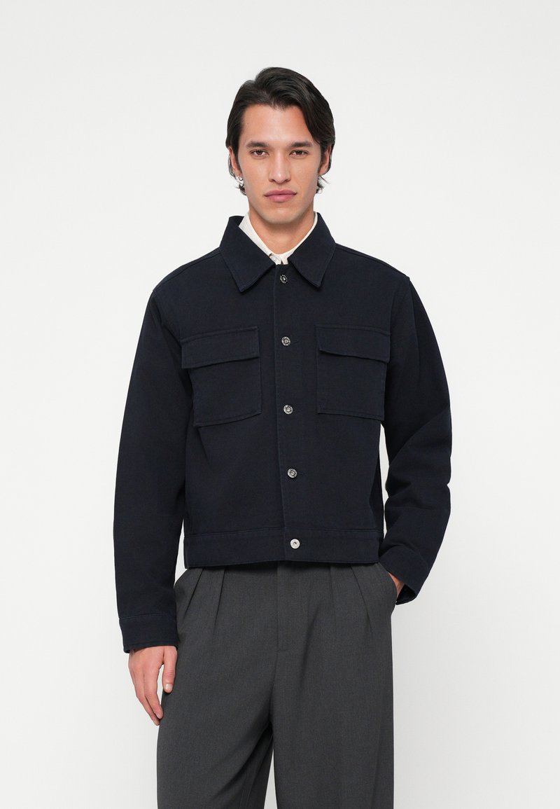 Άνδρας που φοράει μια μαύρη σακάκι με κουμπιά και τσέπες με καπάκι, συνδυασμένο με γκρι φαρδιά παντελόνια, που στέκεται μπροστά σε λευκό φόντο.