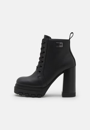 Czarny botek na wysokim obcasie z grubą podeszwą na platformie, sznurowany z przodu, z pętelką do ściągania i metalowym logo Tommy Jeans po boku.
