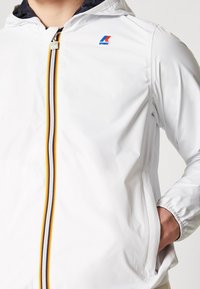 Veste blanche à capuche avec une fermeture éclair rayée jaune, blanche et noire, et un petit logo multicolore sur la poitrine, portée avec une main dans la poche.