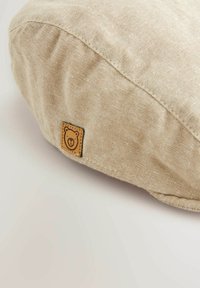 Rundes Kissen aus strukturiertem beigem Stoff mit sichtbaren Nähten. Verfügt über ein kleines, geprägtes Bärenlogo auf einem braunen Patch. Weiches, lässiges Design.
