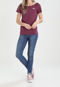 Viininpunainen lyhythihainen t-paita pyöreällä kaula-aukoilla ja logolla vasemmassa rinnassa, yhdistettynä sinisiin denim-farkkuihin ja vaaleanpunaisiin tennareihin.