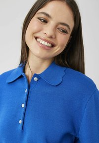 Polo bleu tricoté avec un col festonné, comportant trois boutons et une texture douce. Gros plan sur la partie supérieure du vêtement.