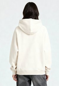 Persoon met lang donker haar die een oversized crèmekleurige hoodie en losse donkergrijze spijkerbroek draagt, gezien van achteren tegen een effen achtergrond.