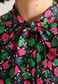 Blusa floreale con uno sfondo scuro, fiori rosa e verdi, un fiocco al collo e una texture di tessuto liscia.