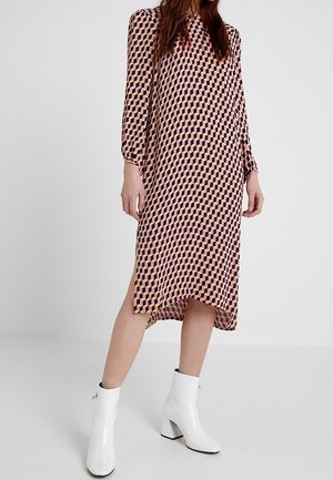 Kobieta w midi sukience z długim rękawem i geometrycznym wzorem, w połączeniu z białymi botkami, stoi na tle jednolitego szarego tła.