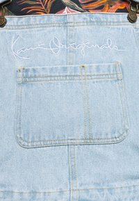 Vaaleansininen farkkiosasto, jossa on suuri tasku, keltaista ommelta, metallisia solkia ja valkoista brodeerattua kursiivitekstiä yläreunan lähellä.