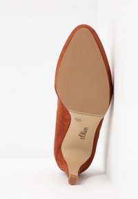 Chaussure à talon haut à bout pointu en suédé marron vue par la semelle, taille 37, marque s.Oliver, placée verticalement contre un coin blanc.