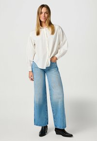 Biała koszula z guzikami o luźnym kroju, z długimi rękawami i zaokrąglonym dołem, zestawiona z jasnoniebieskimi spodniami o szerokich nogawkach i czarnymi botkami za kostkę.