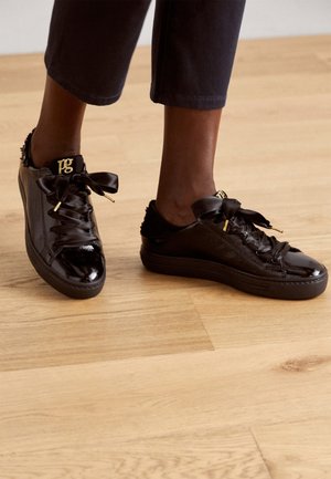 Czarne sneakersy z lakierowanej skóry z szerokimi satynowymi sznurówkami i złotymi elementami, noszone z krótkimi czarnymi spodniami na drewnianej podłodze.