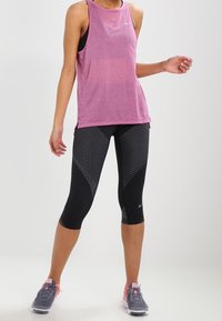 Camiseta deportiva sin mangas rosa con una ligera textura jaspeada, combinada con leggings capri negros con un patrón geométrico. Zapatillas grises con detalles en rosa.
