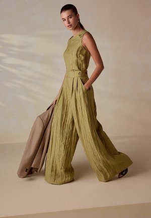 Nainen ilman hihoja olevassa oliivinvihreässä leveälahkeisessa haalariasussa, pitäen beigeä bleiseriä, seisoo beigen taustan edessä, toinen käsi taskussa.