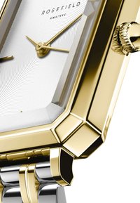 Reloj de metal dorado y plateado con una esfera rectangular, dial con patrón blanco en espiral y corona prominente; cuenta con una caja octagonal y acabado pulido.