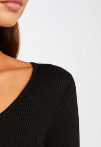 Haut noir côtelé avec un col en V, mettant en valeur un tissu texturé et des bords lisses près de l'épaule. Accent sur le matériau et la structure.