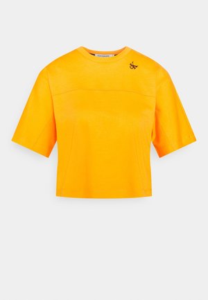 Jasnopomarańczowy skrócony T-shirt z krótkimi rękawami i małym czarnym logo "CK" w pobliżu lewego ramienia.