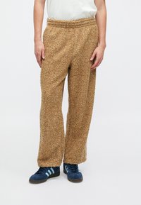 Pantalones de forro polar marrones, esponjosos, con cintura elástica, piernas anchas y una textura suave. Llevados con zapatillas de deporte azul marino con rayas azul claro.
