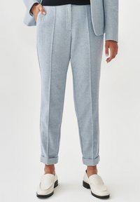 Lichtblauwe op maat gemaakte broek van textuurstof, met plooien, zijzakken en opgerolde boorden. Gecombineerd met crèmekleurige instappers.