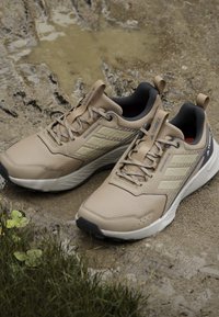 Beige atletiske sko med tekstureret mesh-overdel, forstærket tå og gummisål. Har grå accenter og snørebånd, designet til udendørs brug.