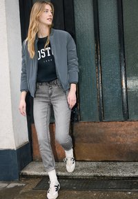 Jeune femme aux longs cheveux blonds debout sur une marche, portant une veste bomber grise, un sweat-shirt noir, un jean gris et des baskets blanches.