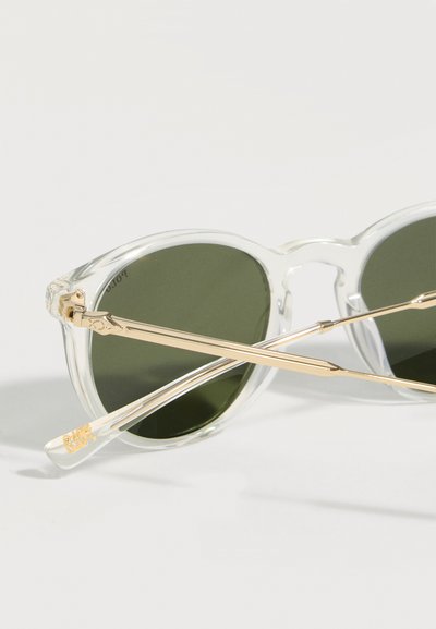 Γυαλιά ηλίου με διαφανές πλαίσιο, στρογγυλούς πράσινους φακούς και χρυσούς μεταλλικούς βραχίονες, τοποθετημένα σε σταυρωτή διάταξη πάνω σε λευκή επιφάνεια.