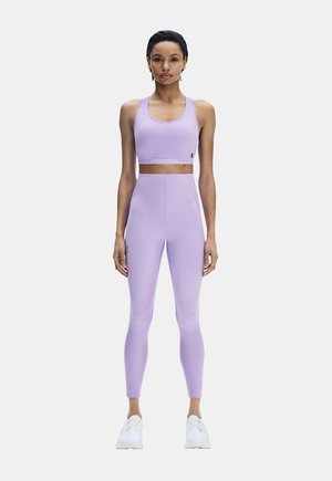 Frau steht, trägt lilafarbenen Sport-BH und passende hoch taillierte Leggings mit weißen Sneakers auf weißem Hintergrund.