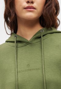 Olivovo zelená mikina s kapucňou so sťahovacou šnúrkou a kovovými okami. Na prednej strane vyšívaný nápis "MSCH COPENHAGEN". Mäkká textúra látky.