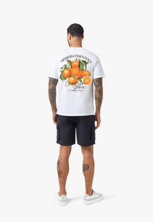 Homme tatoué portant un t-shirt blanc avec un graphisme de fruit orange et le texte « Modern Essentials » au dos, un short noir et des baskets blanches.