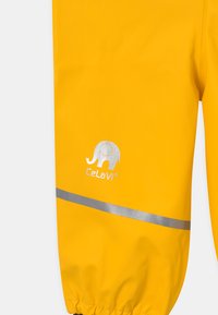 Gule regnbukser i vanntett materiale har et reflekterende sølvbånd og et elefantlogo med merkenavnet "Celavi." Elastiske mansjetter.