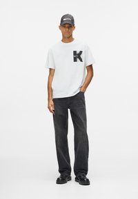 Vīriešu modele, uzvilcis baltu T-kreklu ar lielu melnu "K" grafiku, melnas bikses, melnas kurpes un melnu cepuri, stāv uz viendabīga baltā fona.