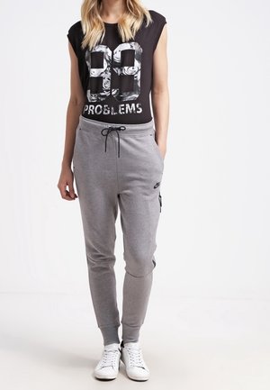 Donna con canotta nera senza maniche con la scritta "99 Problems" e pantaloni della tuta grigi Nike con coulisse, in piedi con le mani nelle tasche.