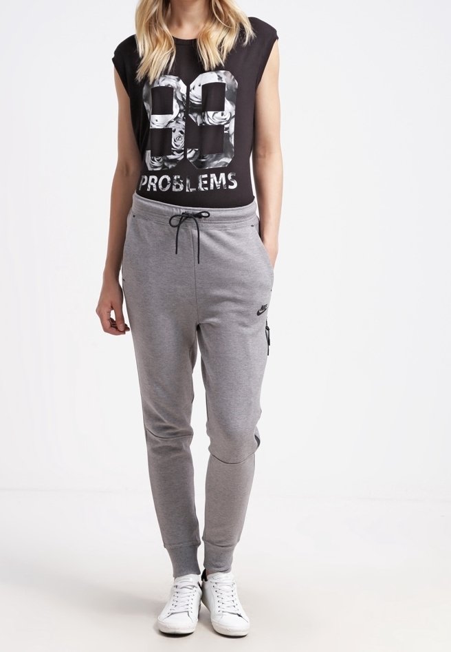 Femme portant un haut noir sans manches avec le texte "99 Problems" et un pantalon de survêtement gris Nike avec cordon de serrage, debout les mains dans les poches.