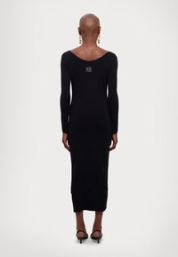 Donna con testa rasata che indossa un lungo abito nero aderente, orecchini a catena d'oro e sandali neri con tacco, in piedi di spalle.