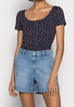 Kobieta ubrana w granatowy top z krótkim rękawem i dekoltem w łódkę, z wzorem w kolorowe posypki, oraz jasnoniebieskie dżinsowe szorty z podwiniętymi nogawkami.