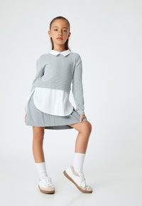 Suéter gris de punto con cuello blanco, superpuesto a una camisa blanca de botones, combinado con una falda plisada gris y zapatillas blancas con acentos beige.