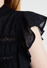 Musta, kevyt paita, jossa on teksturoitu kangas, pyöreitä kuvioita ja röyhelöhihoja. Päällä olevan pitsisomisteen ansiosta vartalon osa lisää visuaalista kiinnostavuutta.