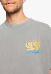 Gray short-sleeve T-shirt featuring a yellow and blue swirl graphic and the text "QUIKSILVER" on the left chest area. Smooth fabric texture.
