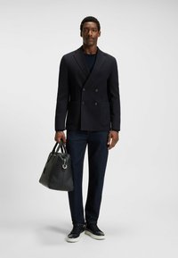 Marinblå dubbelknäppt kavaj i kombination med mörka jeans och svarta sneakers, håller en strukturerad läderväska med handtag.