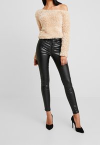 Maglione beige morbido off-the-shoulder abbinato a pantaloni skinny neri lucidi e tacchi a punta neri. Texture morbida e design aderente.