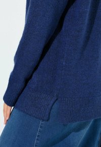 Nahaufnahme einer Person, die einen dunkelblauen Strickpullover mit geripptem Saum und Bündchen trägt, kombiniert mit blauen Jeans.