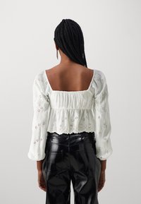 Bílá vyšívaná blůza s čtvercovým výstřihem, dlouhými nabíranými rukávy a stahovaným pasem, spárovaná s černými lesklými texturovanými kalhotami.