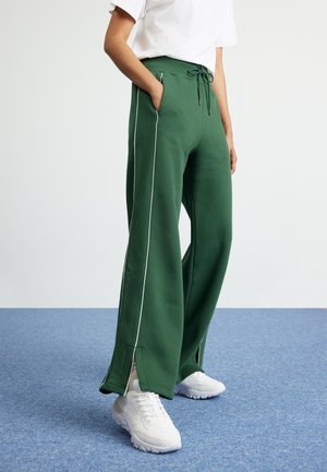 Personne portant un pantalon vert à jambes larges avec un liseré blanc sur les côtés et des fentes à l'avant, associé à des baskets blanches et un t-shirt blanc.
