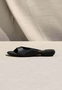 Μαύρο σανδάλι slip-on με ανοιχτό δάχτυλο και υφασμένο πάτο που ακουμπά σε ανοικτό μπεζ επιφάνεια, με ουδέτερο υφασμάτινο φόντο.