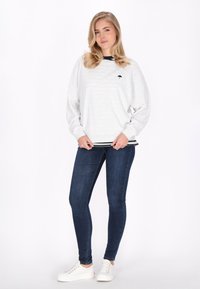 Felpa grigia chiara con colletto nero e un motivo discreto, caratterizzata da un piccolo logo nero sul petto. Abbinata a jeans skinny blu scuro.