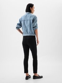 Sieviete stāv ar muguru, ģērbta gaiši zilā denima jakā, melnās biksēs un melnos loafers uz vienkārša balta fona.