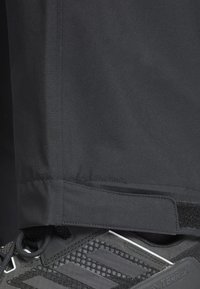 Pantalons étanches noirs avec une texture lisse, dotés d'une fermeture à Velcro à la cheville, associés à une chaussure de sport noire avec un dessus texturé.