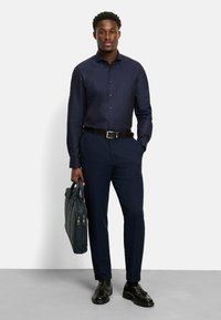 Navyblau gemusterte Hemdbluse, navyblaue Hosen, brauer Gürtel, schwarze Lederschuhe, hält eine dunkelgraue Lederaktentasche.