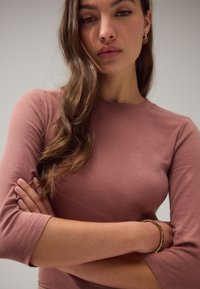 Pikad varrukad, soonikkoes topp tuhmroosas toonis, ümmarguse kaelusega ja keha järgiva lõikega. Kandmisel on käevõru ja detailselt kujundatud kõrvarõngad.