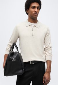 Ein Mann trägt ein hellbeiges Langarm-Poloshirt und schwarze Hosen und hält eine schwarze Ledertasche mit Griffen vor einem einfarbigen Hintergrund.