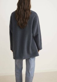 Giacca in fleece grigio scuro con una vestibilità rilassata, dotata di maniche lunghe, orlo dritto e una superficie morbida e testurizzata. Indossata sopra pantaloni azzurri chiari.