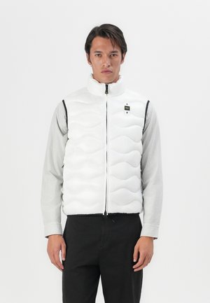 White puffer vest with a high collar and wave pattern. Features a front zipper and black accents. Model wears a striped shirt underneath.