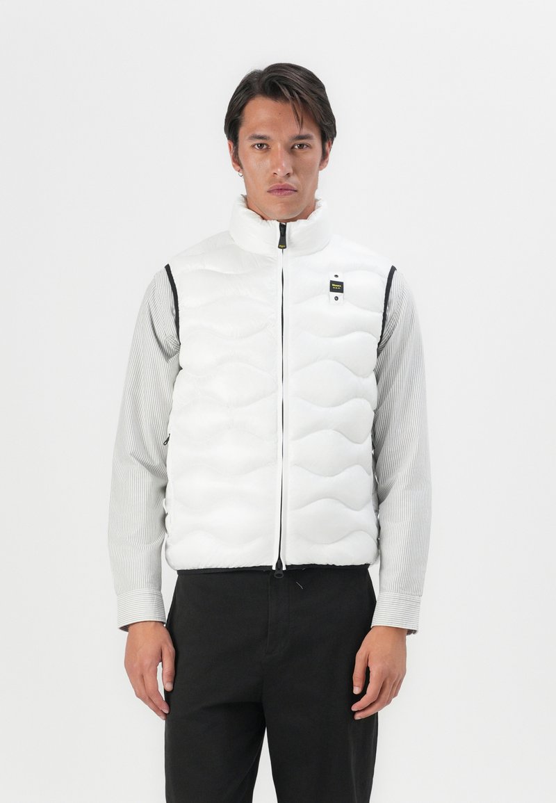 White puffer vest with a high collar and wave pattern. Features a front zipper and black accents. Model wears a striped shirt underneath.